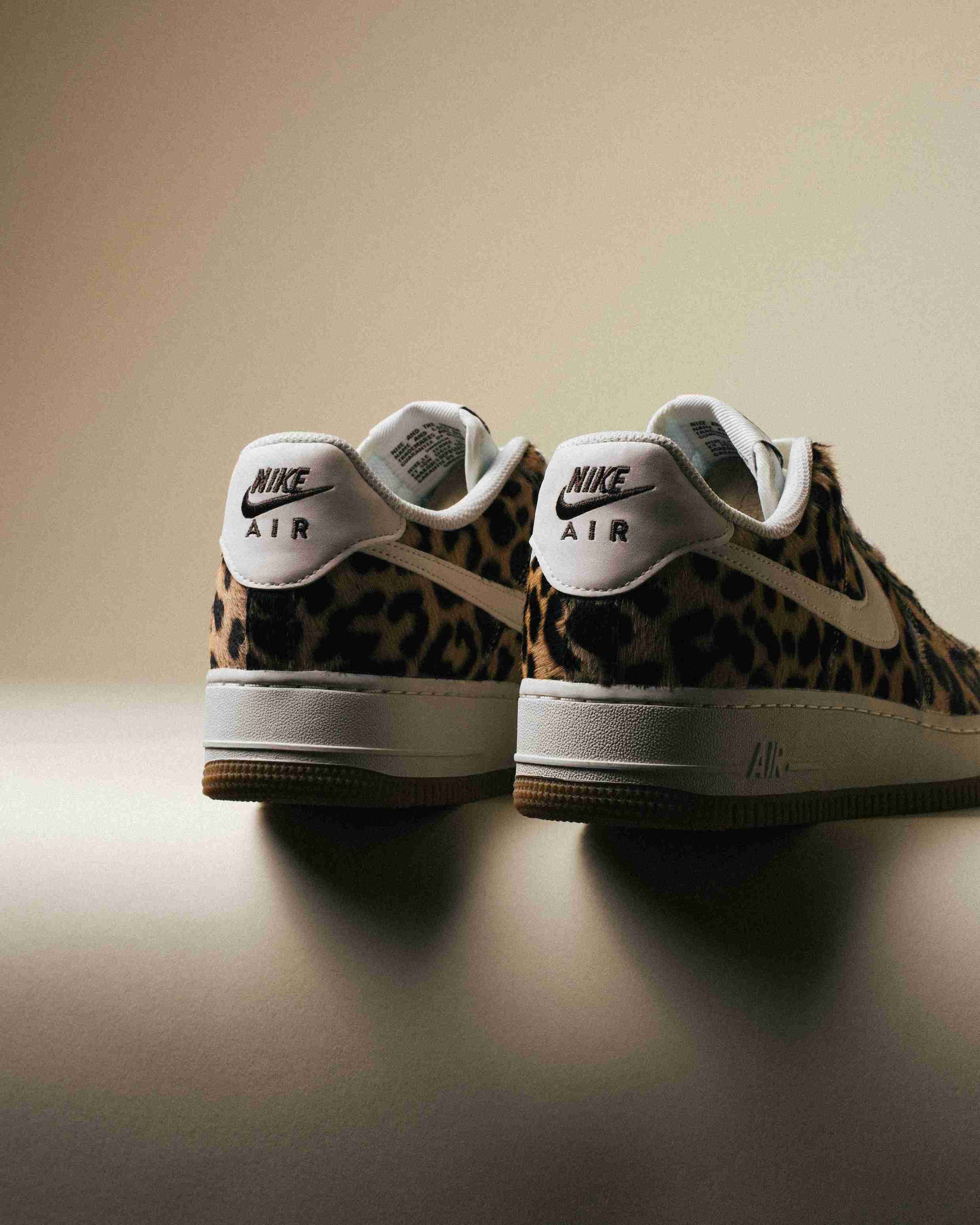 Paire de sneakers Nike Air Force 1 à motif léopard, placée à côté d’une boîte Nike grise sur un fond beige, vue artistique avec lumière douce.